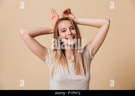 Foto di adorabile donna europea in t-shirt casual sorridente e mostrando bunny orecchie al suo capo isolate su sfondo beige Foto Stock