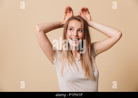 Foto di divertenti affascinante ragazza 20s in t-shirt casual sorridente e mostra orecchie di coniglio al suo capo isolate su sfondo beige Foto Stock