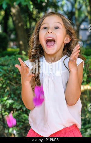 Carino cinque anni bambina con gli occhi blu e i capelli biondi che mostra la felicità e la sorpresa sul suo viso con un parco come sfondo Foto Stock