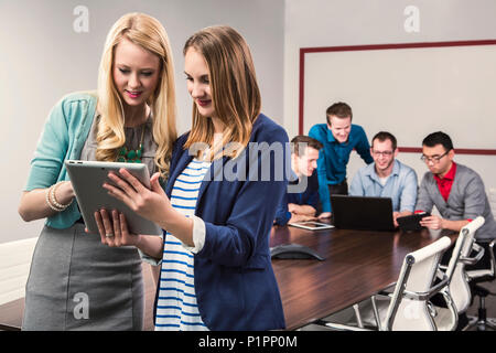 Due giovani millenario professionisti aziendali guardando un tablet mentre si lavora insieme in una sala conferenze con i loro coetanei Foto Stock