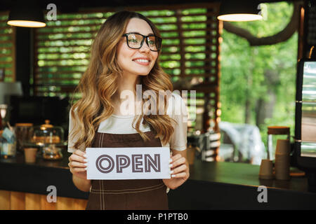 Ritratto di un attraente giovane barista ragazza nel grembiule tenendo aperta la scheda di segno permanente, mentre presso il cafe Foto Stock