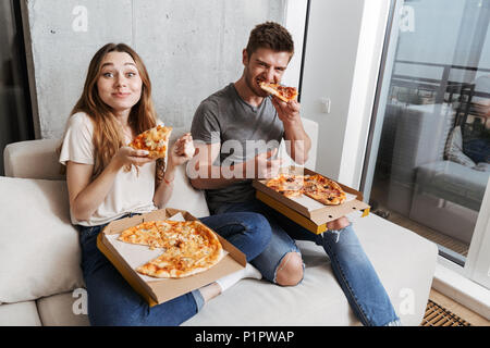Soddisfatto coppia giovane mangiare la pizza seduti insieme sul lettino a casa Foto Stock