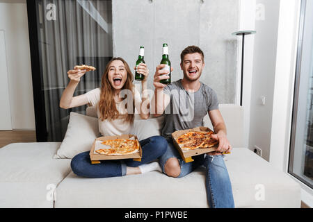 Allegro coppia giovane di mangiare pizza e bere birra seduti insieme sul lettino a casa e guardare la TV Foto Stock