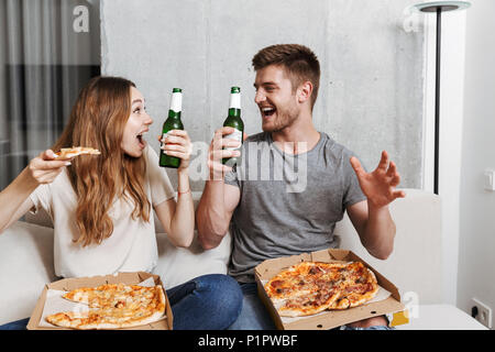 Gioiosa coppia giovane di mangiare pizza e bere birra seduti insieme sul lettino a casa e guardare la TV Foto Stock