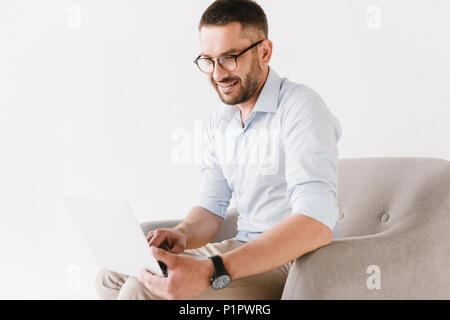 Ritratto orizzontale del barbuto caucasian ragazzo indossa eleganti abiti formali seduto in poltrona e di lavoro sul computer portatile isolato su sfondo bianco Foto Stock