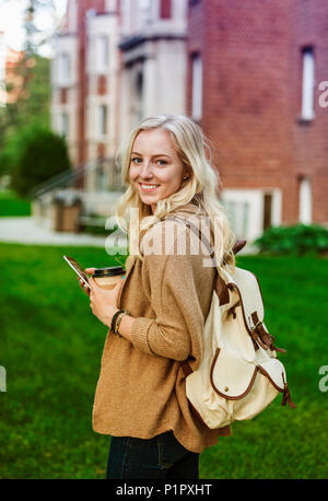 Una bella giovane donna con lunghi capelli biondi tenendo una tazza di caffè e testando sul suo smartphone mentre si cammina in un campus universitario si ferma a posare... Foto Stock