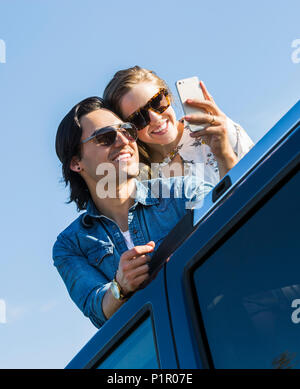 Una giovane coppia stand up attraverso il tettuccio apribile del loro veicolo e prendere un autoritratto con il loro telefono cellulare; Edmonton, Alberta, Canada Foto Stock