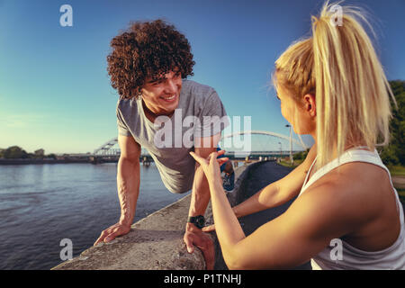 Giovane coppia fitness facendo allenamento sulla parete lungo il fiume in un tramonto. L'uomo è accovacciato e azienda kettlebell e la donna lo appoggio. Foto Stock