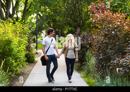Una giovane coppia a piedi giù per un sentiero sul campus universitario tenendo le mani e parlando; Edmonton, Alberta, Canada Foto Stock