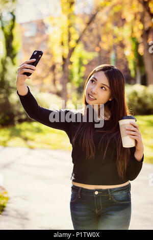 Una giovane donna internazionale studente universitario sta tenendo una tazza di caffè e pone per un autoritratto con il suo smart phone Foto Stock