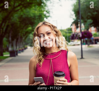 Una giovane donna guarda la fotocamera mentre si cammina in una strada con un caffè vicino a un campus universitario e i messaggi di testo sul suo smart phone Foto Stock