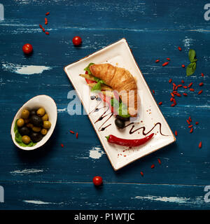 Appetitosi sandwich vegetariano con croissant e verdure giacente sulla piastra piana di forma rettangolare, ciotola di olive e pomodori ciliegia ed essiccati goji bacche sc Foto Stock