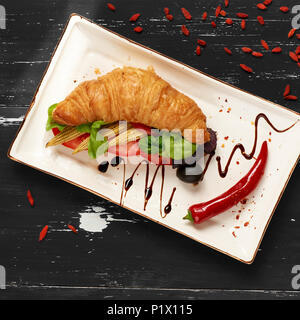 Appetitosi sandwich vegetariano con croissant e verdure giacente sulla piastra piana di forma rettangolare, ciotola di olive e pomodori ciliegia ed essiccati goji bacche sc Foto Stock