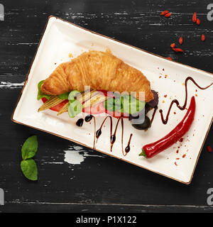 Appetitosi sandwich vegetariano con croissant e verdure giacente sulla piastra piana di forma rettangolare, ciotola di olive e pomodori ciliegia ed essiccati goji bacche sc Foto Stock