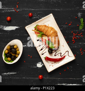 Appetitosi sandwich vegetariano con croissant e verdure giacente sulla piastra piana di forma rettangolare, ciotola di olive e pomodori ciliegia ed essiccati goji bacche sc Foto Stock
