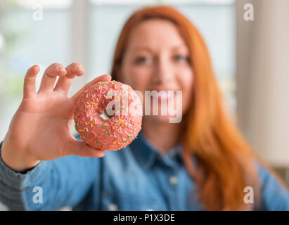 Redhead donna ciambella di contenimento a casa con una faccia felice in piedi e sorridente con un sorriso sicuro che mostra i denti Foto Stock