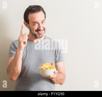 Senior uomo mangiare patatine sorpreso con un'idea o domanda puntare il dito con la faccia felice, numero uno Foto Stock