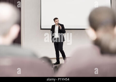 Altoparlante conduce il business della conferenza in piedi di fronte a un grande schermo bianco sul palco in una sala conferenze Foto Stock