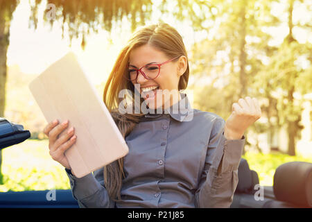 Allegra donna in piedi con la sua nuova vettura la ricerca di lavoro con computer di pad in un urbano d'estate il parco Foto Stock