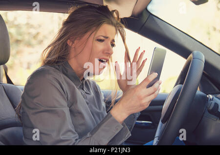 Giovani arrabbiati ragazza utilizzando smartphon con espressione di rabbia e di ansia mentre è seduto all'interno e la guida auto. Foto Stock