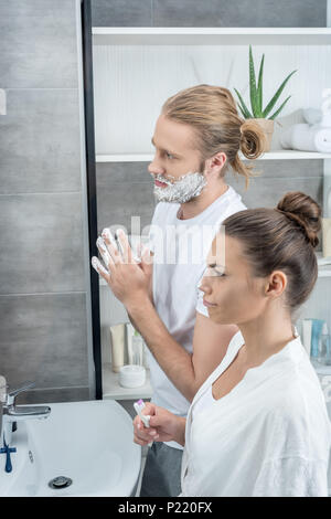 L'uomo la rasatura la sua barba mentre donna spazzolare i denti in bagno al mattino Foto Stock