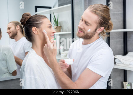 Giovani barbuto marito l'applicazione di crema per il viso sulla guancia della moglie nella stanza da bagno Foto Stock