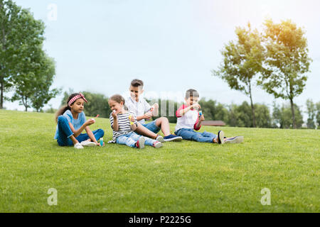 Adorabili bambini multietnica soffiando bolle di sapone seduti insieme in posizione di parcheggio Foto Stock