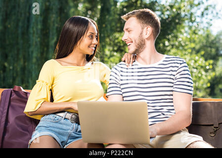 Felice coppia multietnica tramite computer portatile mentre è seduto sulla panchina nel parco e sorridente ogni altro Foto Stock