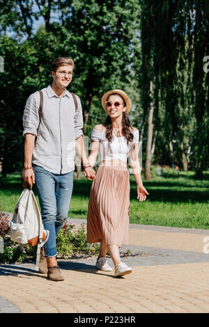 Elegante coppia multiculturale tenendo le mani mentre passeggiate nel parco di estate insieme Foto Stock