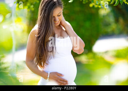 Sorridente donna incinta outdoor permanente tenendo le mani sulla pancia Foto Stock