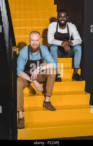 Due multietnica sorridente proprietari dei coffee shop in grembiuli seduti sulle scale Foto Stock