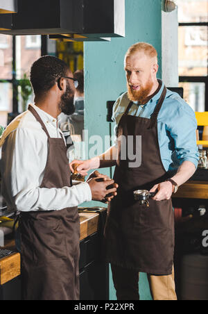 Giovane maschio multietnica proprietari dei coffee shop in grembiuli parlando in coffee shop Foto Stock