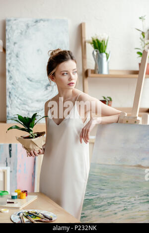 Bella giovane artista con verde pianta in vaso appoggiandosi a immagine sul cavalletto e guardando lontano in art studio Foto Stock