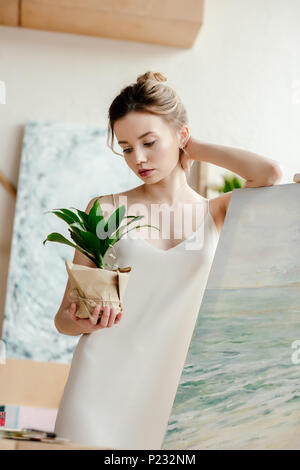 Bella giovane artista appoggiata al foto sul cavalletto e guardando il verde pianta in vaso Foto Stock