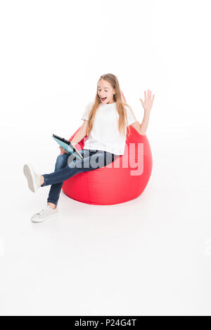 Excited donna giovane con tavoletta digitale e seduta in rosso poltrona sacco, isolato su bianco Foto Stock