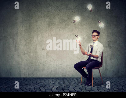 Felice smart uomo seduto su una sedia contro il grigio muro di cemento e giocoleria con lampadine avente molte idee brillanti. Foto Stock