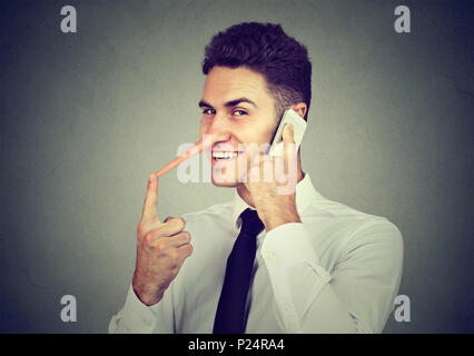 Sly giovane con il naso lungo parlando al telefono cellulare isolata sul muro grigio Sfondo. Concetto bugiardo. Emozione umana sentimenti, tratti caratteriali Foto Stock