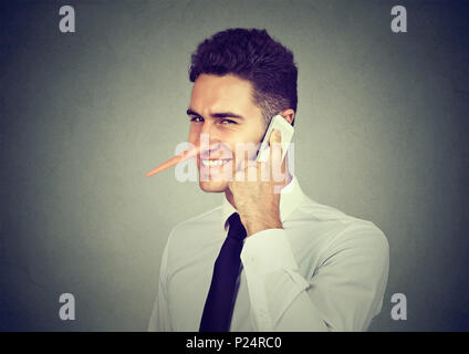 Sly giovane con il naso lungo parlando al telefono cellulare isolata sul muro grigio Sfondo. Concetto bugiardo. Emozione umana sentimenti, tratti caratteriali Foto Stock