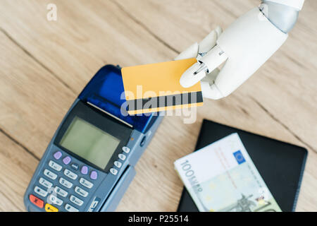 Vista ravvicinata di mano del robot holding credito carta al di sopra del denaro e il terminale di pagamento sul tavolo di legno Foto Stock