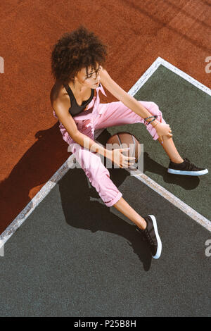 Giovane africano-donna americana nel reggiseno sportivo e tute rosa seduta sul terreno del campo sportivo con campi da basket ball Foto Stock