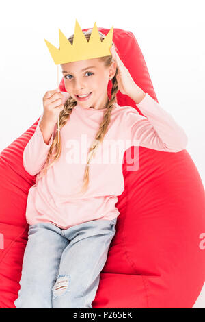 Bambino sorridente con la corona di cartone seduto su una red bean bag, isolato su bianco Foto Stock