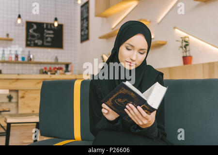 Concentrato di bella donna musulmana leggere il Corano in cafe Foto Stock