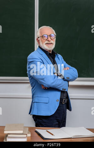 Sorridente i capelli grigi professore in piedi con le braccia incrociate Foto Stock