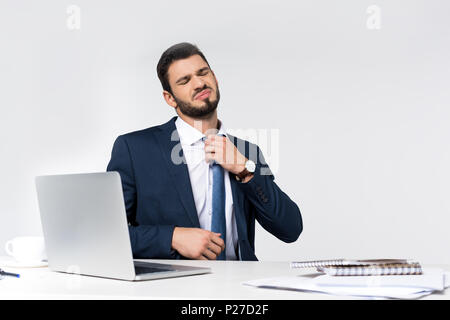 Giovane imprenditore oberato di lavoro con gli occhi chiusi cravatta regolazione stando seduti al posto di lavoro Foto Stock