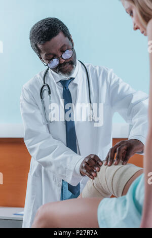 African American medico mettendo sul bendaggio elastico sul ginocchio del paziente Foto Stock