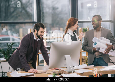 Multietnica sorridente gli architetti che lavorano con blueprint e computer in ufficio moderno Foto Stock