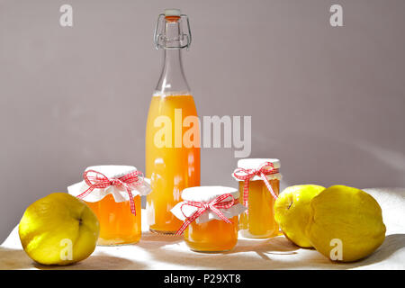 In casa cotognata e succo in un biberon e vasetti di vetro con mele cotogne su una tovaglia di lino sotto il sole di fronte a uno sfondo neutro. Foto Stock