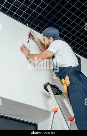 Basso angolo vista del giovane elettricista in piedi sulla scaletta e fissaggio allarme home Foto Stock