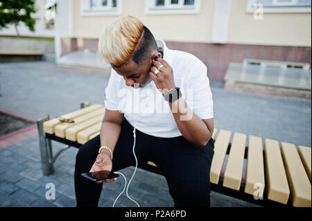 Elegante arabian ragazzo musulmano con capelli originariamente poste sulle strade, seduti su una panchina e ascoltare musica dagli auricolari del telefono. Foto Stock