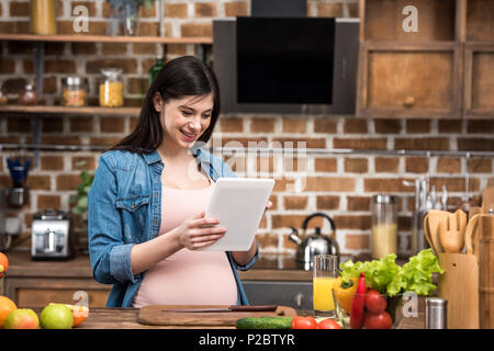 Sorridente giovane donna incinta con tavoletta digitale durante la cottura in cucina Foto Stock